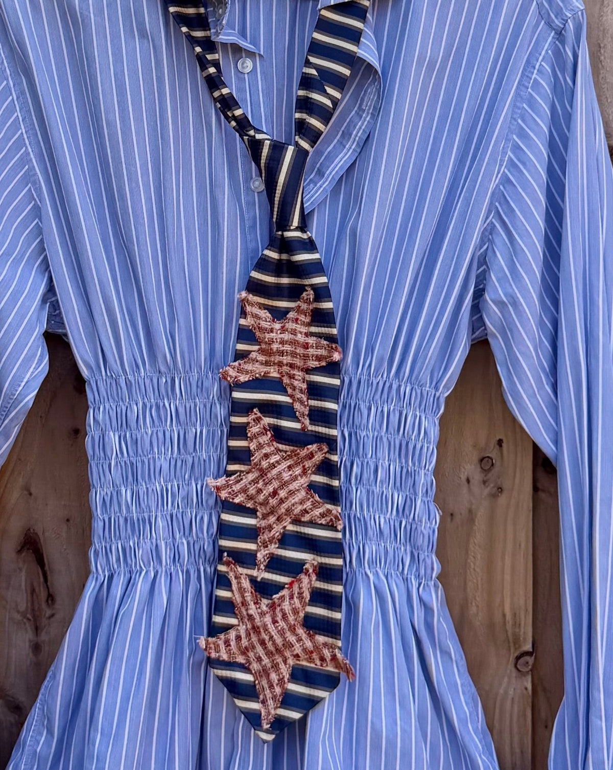 A light blue and white vertically striped long-sleeve blouse with a smocked waist and a decorative embroidered tie detail, hanging on a wooden fence outdoors.