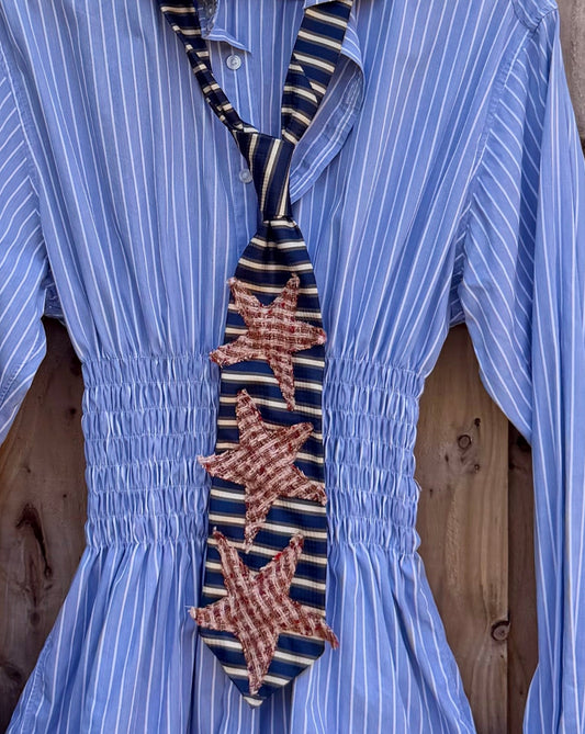 A light blue and white vertically striped long-sleeve blouse with a smocked waist and a decorative embroidered tie detail, hanging on a wooden fence outdoors.