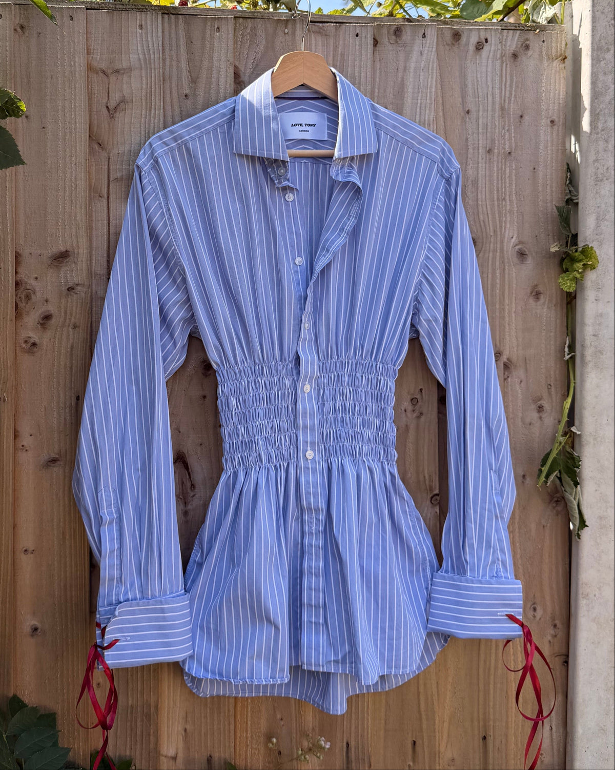 A light blue and white vertically striped long-sleeve blouse with a smocked waist and red tie details on the cuffs, hanging on a wooden hanger against a wooden fence with green foliage.