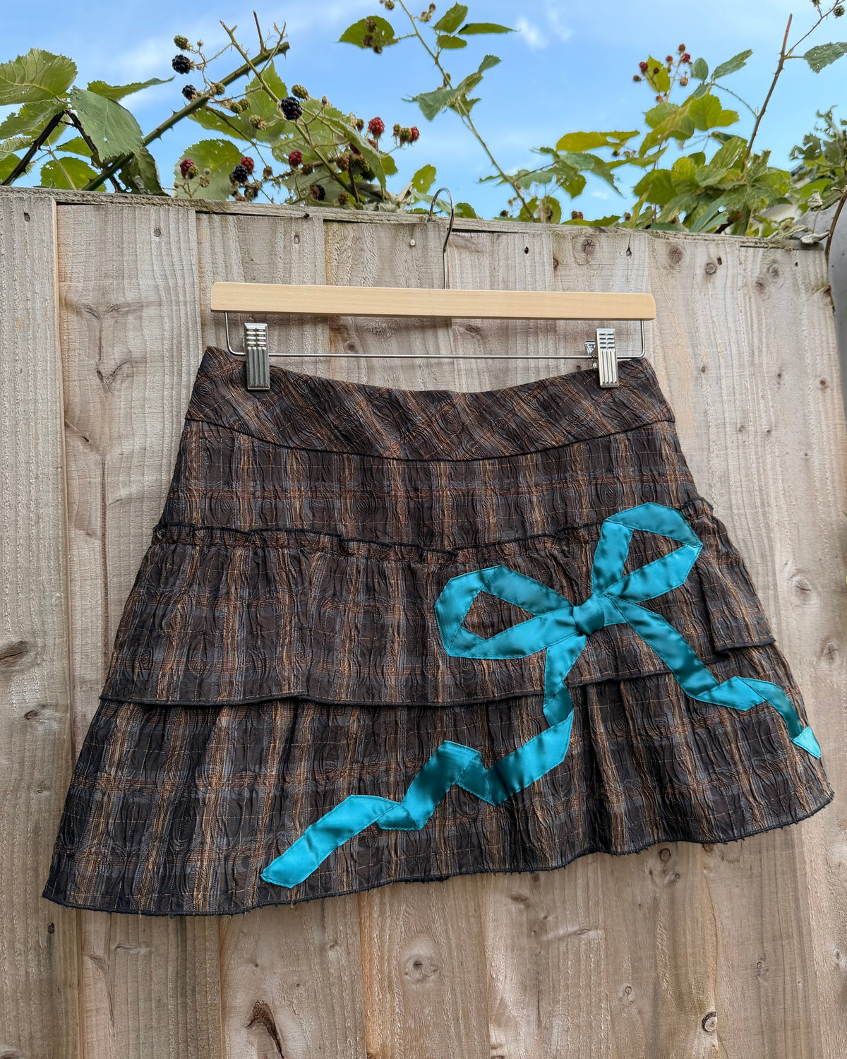 A tiered mini skirt with a brown and black plaid pattern, featuring a large turquoise ribbon bow and matching ribbon draped across the front, hanging from a wooden hanger on a wooden fence outdoors.