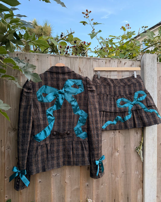 A matching plaid jacket and skirt set hanging on a wooden fence outdoors. The dark brown and black plaid fabric features large bright turquoise ribbon bows on the back of the jacket and on the skirt, with matching ribbon bows tied at the cuffs of the jacket.