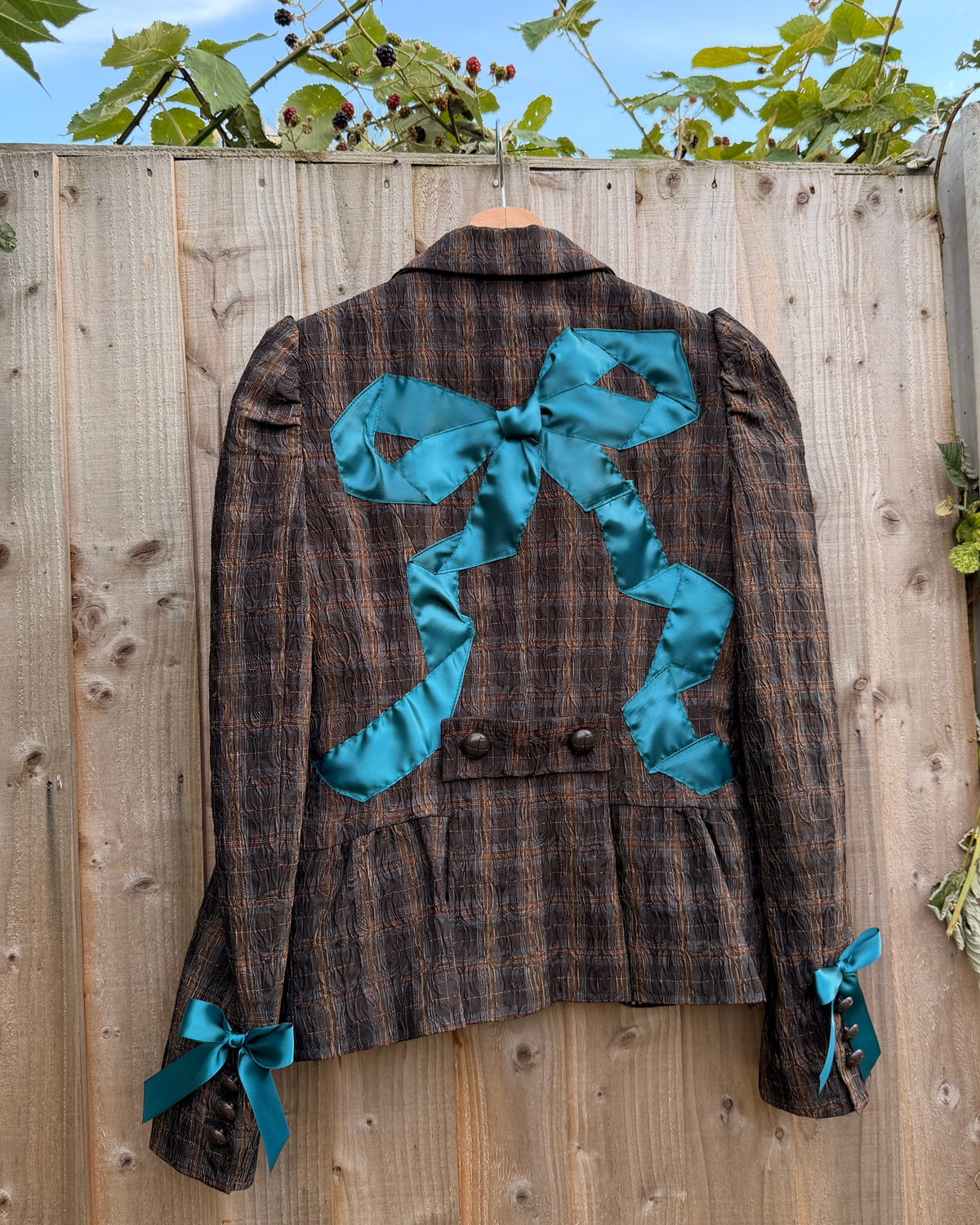 A brown and black plaid cropped jacket with a large teal satin bow on the chest and matching teal bow ties at the cuffs, laid flat on a wooden fence outdoors.